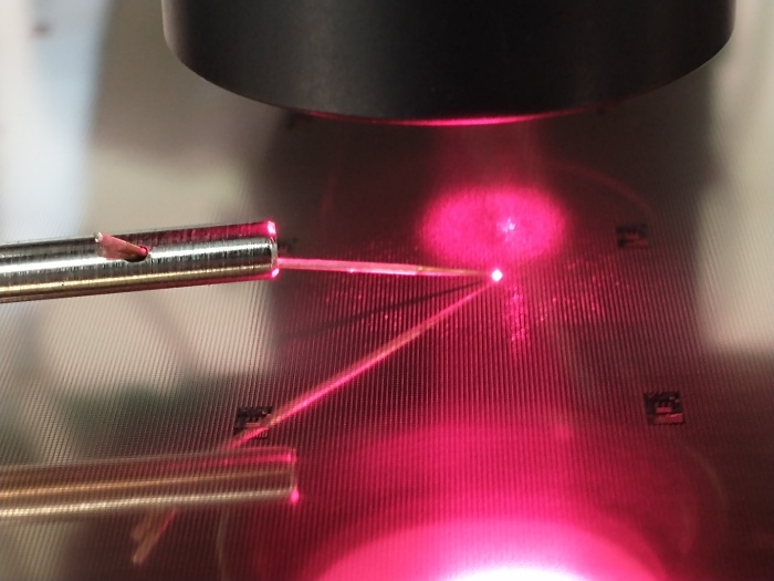 Probe needles contacting a VCSEL die under a microscope, with the laser spot visible on the wafer surface.